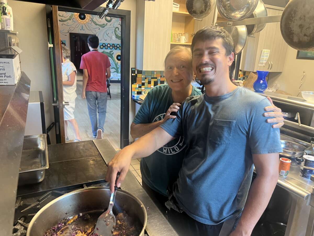 Deux personnes posent par un poêle alors que l'on mélange des aliments dans un pot. 